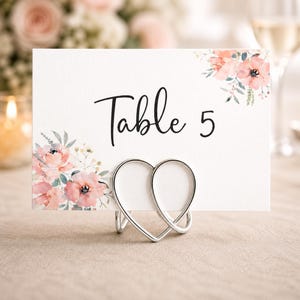 May include: A white table number card with the word "Table" and the number "5" in black script. The card is decorated with watercolor floral accents in pink and green. It is held by a silver heart-shaped holder.