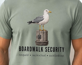 May include: A sage green t-shirt featuring a graphic of a seagull standing on a wooden piling wrapped in rope. Below the graphic, the text reads BOARDWALK SECURITY followed by the words unpaid • uninvited • unblinking in black lettering.