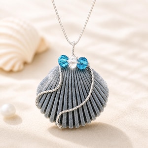May include: A seashell pendant necklace with a silver chain. The pendant is a gray scallop shell adorned with blue and clear crystal beads. The necklace is displayed on a sandy surface, with a seashell and a pearl in the background.