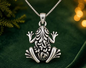May include: A silver-toned metal pendant shaped like a frog with intricate embossed detailing on its back and limbs, suspended from a delicate silver-toned box chain necklace. The frog figure features large, circular eyes and splayed feet.