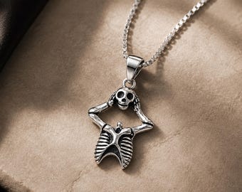 May include: A silver-toned metal pendant necklace featuring a stylized skeleton figure with its hands placed on its head. The pendant is suspended from a delicate box chain, showcasing oxidized detailing that highlights the ribcage and skull.