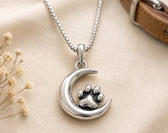 May include: A silver-toned metal pendant necklace featuring a crescent moon shape with a raised paw print design in the center. The pendant hangs from a thin, square-link chain, showcasing a polished finish with dark recessed detailing.