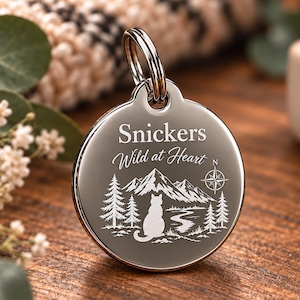 May include: A circular silver-toned metal pet tag featuring an engraved design of a cat sitting before mountains, pine trees, and a winding path. The text reads Snickers and Wild at Heart, accompanied by a compass rose symbol and a metal split ring.