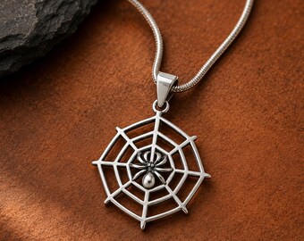 May include: A silver-toned pendant necklace featuring a circular spider web design with a small spider positioned in the center. The pendant is attached to a thin, flexible snake chain, showcasing a polished metallic finish and intricate detailing.