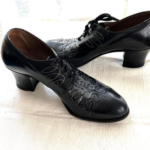 May include: A pair of black leather heeled shoes with lace-up closures and intricate floral designs. The shoes have a low block heel and a rounded toe.