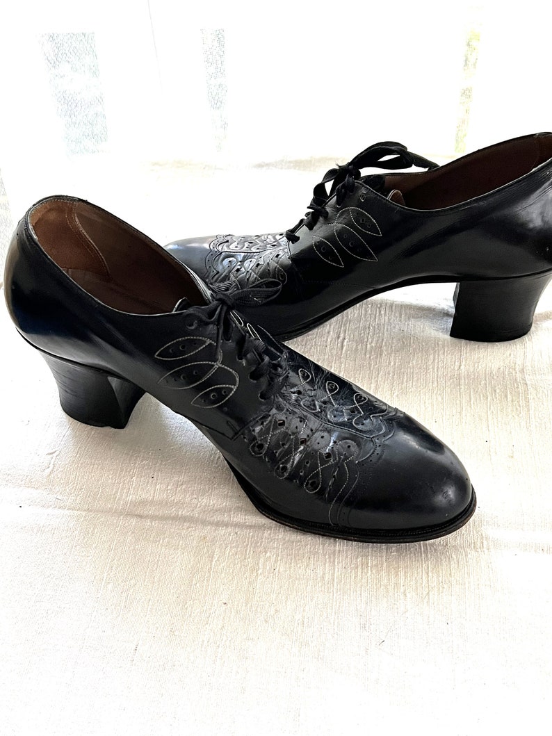 May include: A pair of black leather heeled shoes with lace-up closures and intricate floral designs. The shoes have a low block heel and a rounded toe.