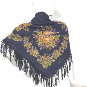 May include: Navy blue square scarf with a floral pattern in shades of yellow, green, and pink. The scarf has a fringe trim in black. The scarf is draped over a mannequin.