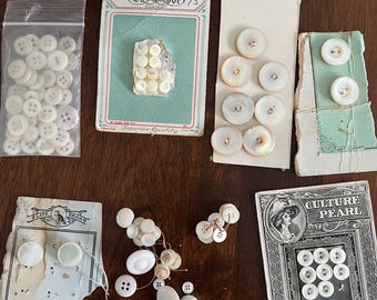 Op de afbeelding: Een assortiment witte parelmoerknopen in verschillende maten en stijlen, sommige in de originele verpakking. De verpakkingen bevatten de merken "La Mode", "Elite Brand" en "Culture Pearl".