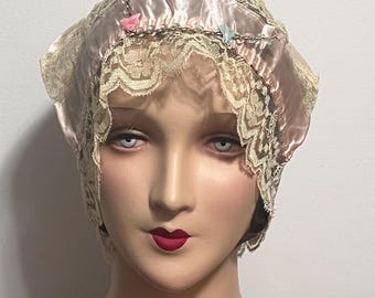 May include: A vintage-style bonnet in soft pink satin, adorned with delicate beige lace trim. The bonnet features small, colorful accents and a chain detail. The head covering is displayed on a mannequin head.