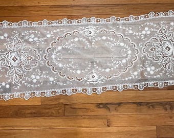May include: White lace table runner with a delicate floral pattern. The runner has a scalloped edge and is perfect for adding a touch of elegance to any table setting.