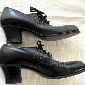 May include: A pair of black leather heeled shoes with a lace-up closure and a decorative leaf design. The shoes have a chunky heel and a rounded toe.