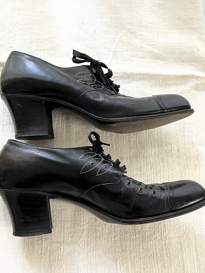 May include: A pair of black leather heeled shoes with a lace-up closure and a decorative leaf design. The shoes have a chunky heel and a rounded toe.