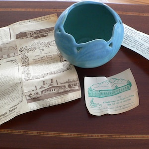 Op de afbeelding: Een lichtblauwe keramische pot met een uniek ontwerp. De pot heeft een brede opening en een ronde bodem. De pot staat op een houten oppervlak. De tekst "Van Briggle Pottery" is zichtbaar in de afbeelding.