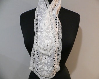 May include: A white lace cravat with a delicate floral pattern. The cravat is tied in a knot and is displayed on a black mannequin.