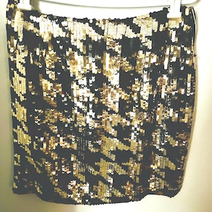 May include: A black sequin mini skirt with gold and bronze sequins. The skirt is hanging on a white hanger. The sequins are densely packed, creating a shimmering effect. The skirt is a straight, fitted style.