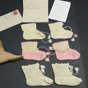 Op de afbeelding: Een assortiment vintage babyslofjes in crème en roze, met bijpassende kwastjes. Een houten schoenleest en handgeschreven brieven zijn ook aanwezig. De slofjes zijn waarschijnlijk met de hand gebreid of gehaakt, met een delicaat, open patroon.
