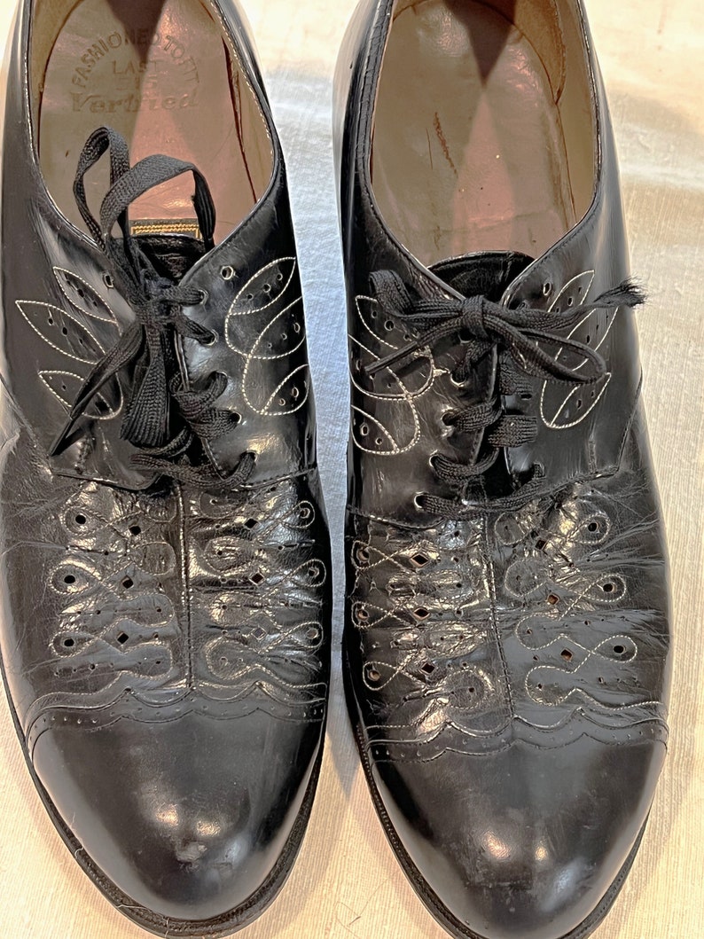 May include: A pair of black leather lace-up shoes with decorative stitching. The shoes have a pointed toe and a low heel. The brand name "Fashion Craft" and the text "Last Variflex" are visible on the inside of the shoes.