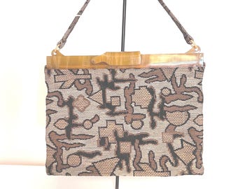 May include: A vintage handbag with a geometric pattern in shades of brown and beige. The bag features a light brown handle and a clasp. The bag is displayed on a black metal stand against a white background.