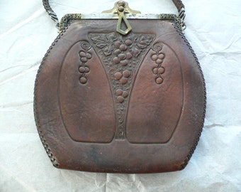 May include: A brown leather purse with a metal clasp and a decorative grape design. The purse has a long strap.