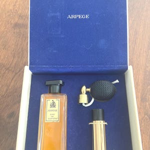 Puede incluir: Un juego de perfumes vintage en una caja azul. El juego incluye un frasco de perfume rectangular con tapa negra, un atomizador dorado y un pequeño recipiente dorado. El frasco de perfume tiene una etiqueta que dice "Arpege Eau de Lanvin".