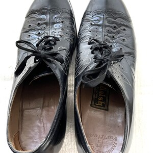 May include: A pair of black leather lace-up shoes with a decorative pattern on the toe. The shoes have a label that reads "Fashioned to Last 515  Very  Feel  Well-ish Boston".