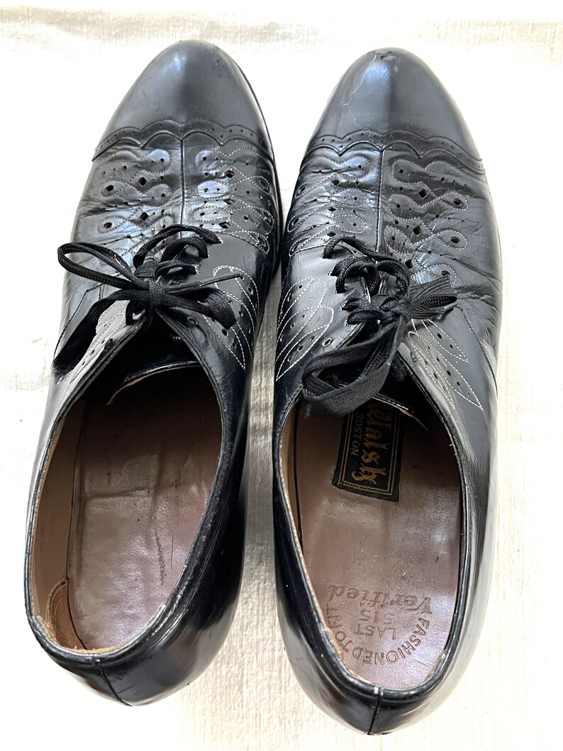 May include: A pair of black leather lace-up shoes with a decorative pattern on the toe. The shoes have a label that reads "Fashioned to Last 515  Very  Feel  Well-ish Boston".