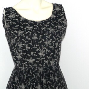 Può includere: Abito nero senza maniche con motivo floreale. L'abito presenta uno scollo tondo e un corpetto aderente che si allarga in vita. Il motivo floreale è di colore beige chiaro, creando un contrasto con il tessuto nero.