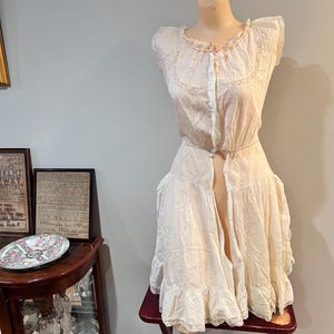 May include: Cream-colored vintage dress displayed on a mannequin. The dress features delicate embroidery around the neckline and sleeves, with a button-down front and a ruffled hem. The dress is displayed on a small, red wooden stool.