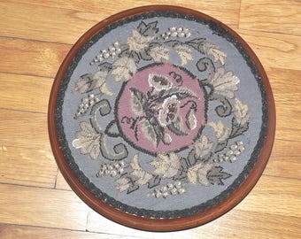 May include: A round, wooden framed needlepoint with a floral design in shades of grey, white, and pink. The design features a central motif of a flower with leaves and vines.