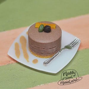 Puede incluir: Un pastel de chocolate en miniatura con una cobertura decorativa de cáscara de naranja confitada, un pistacho y un trozo de chocolate negro. El pastel está sobre un plato blanco con un tenedor plateado. El plato tiene un diseño decorativo.