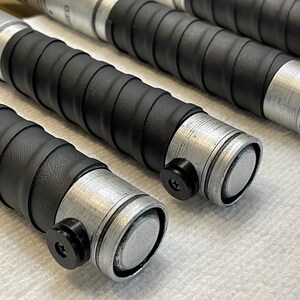 K&ouml;nnte beinhalten: Nahaufnahme von vier schwarzen und silbernen Lichtschwertern mit einem strukturierten Griff. Die Griffe haben eine silberne Metallkappe und einen schwarzen Knopf.