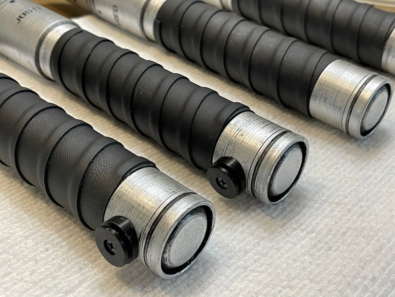 K&ouml;nnte beinhalten: Nahaufnahme von vier schwarzen und silbernen Lichtschwertern mit einem strukturierten Griff. Die Griffe haben eine silberne Metallkappe und einen schwarzen Knopf.