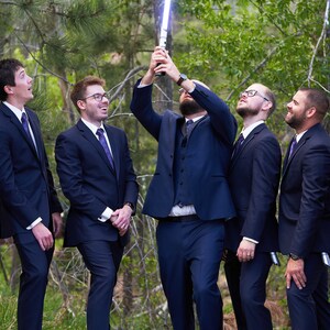 K&ouml;nnte beinhalten: Sechs M&auml;nner in dunkelblauen Anz&uuml;gen und violetten Krawatten stehen in einer Reihe und schauen zu einem leuchtenden blauen Lichtschwert auf, das der Mann in der Mitte h&auml;lt.