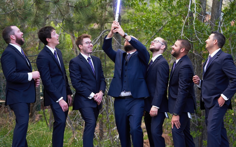 K&ouml;nnte beinhalten: Sechs M&auml;nner in dunkelblauen Anz&uuml;gen und violetten Krawatten stehen in einer Reihe und schauen zu einem leuchtenden blauen Lichtschwert auf, das der Mann in der Mitte h&auml;lt.