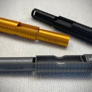 Puede incluir: Tres piezas impresas en 3D en oro, negro y gris. Cada pieza es un cilindro hueco con una ranura que recorre su longitud. Las piezas están etiquetadas como "mavlabs 3D".