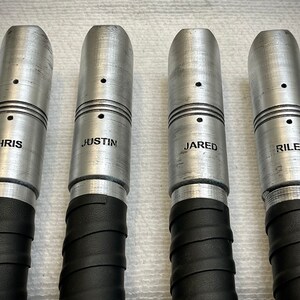 K&ouml;nnte beinhalten: Vier silberne Metallr&ouml;hren mit schwarzem Klebeband um den Boden gewickelt. Jede R&ouml;hre hat einen kleinen schwarzen Punkt und einen eingravierten Namen: Chris, Justin, Jared und Riley.