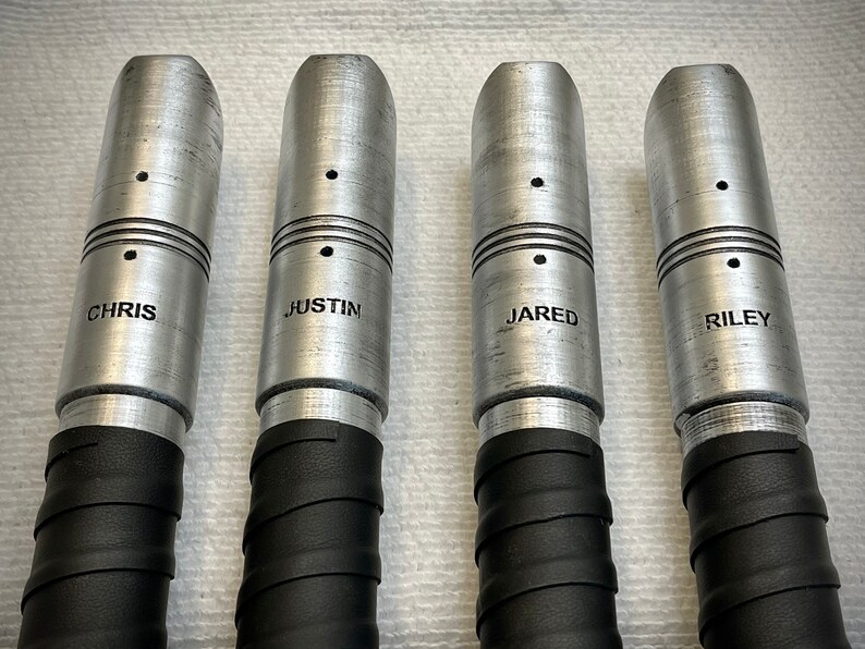 K&ouml;nnte beinhalten: Vier silberne Metallr&ouml;hren mit schwarzem Klebeband um den Boden gewickelt. Jede R&ouml;hre hat einen kleinen schwarzen Punkt und einen eingravierten Namen: Chris, Justin, Jared und Riley.