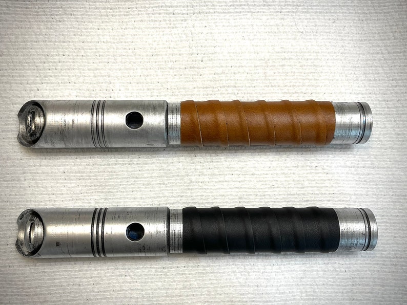 K&ouml;nnte beinhalten: Zwei Lichtschwertgriffe, einer mit einer braunen Lederummantelung und der andere mit einer schwarzen Lederummantelung. Beide Griffe sind aus Metall und haben eine silberne Oberfl&auml;che.