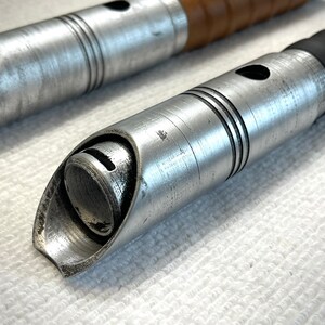 K&ouml;nnte beinhalten: Nahaufnahme der silbernen Metallspitze eines Lichtschwertgriffs. Der Griff hat eine strukturierte Oberfl&auml;che und einen schwarzen Gummibeschlag.