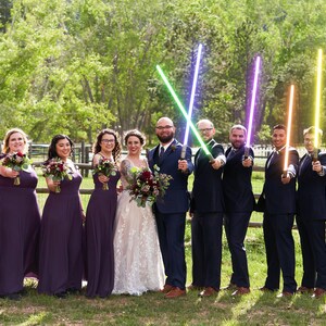 K&ouml;nnte beinhalten: Eine Hochzeitsgesellschaft posiert f&uuml;r ein Foto, die Trauzeugen halten bunte Lichtschwerter. Die Brautjungfern tragen lila Kleider und halten Blumenstr&auml;u&szlig;e. Braut und Br&auml;utigam stehen im Zentrum, der Br&auml;utigam h&auml;lt ein gr&uuml;nes Lichtschwert und die Braut einen Strau&szlig; roter Blumen.