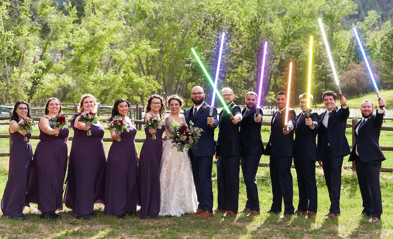K&ouml;nnte beinhalten: Eine Hochzeitsgesellschaft posiert f&uuml;r ein Foto, die Trauzeugen halten bunte Lichtschwerter. Die Brautjungfern tragen lila Kleider und halten Blumenstr&auml;u&szlig;e. Braut und Br&auml;utigam stehen im Zentrum, der Br&auml;utigam h&auml;lt ein gr&uuml;nes Lichtschwert und die Braut einen Strau&szlig; roter Blumen.