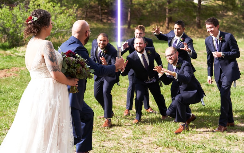 K&ouml;nnte beinhalten: Eine Hochzeitsgesellschaft posiert f&uuml;r ein Foto, wobei der Br&auml;utigam ein blaues Lichtschwert h&auml;lt. Das Brautpaar steht im Vordergrund, w&auml;hrend die Trauzeugen im Hintergrund sind. Die Trauzeugen tragen alle dunkle Anz&uuml;ge und machen spielerische Posen.