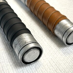 K&ouml;nnte beinhalten: Zwei Lichtschwerter mit Griffen in verschiedenen Farben. Ein Griff hat einen schwarzen Ledergriff und der andere einen braunen Ledergriff. Beide Griffe haben eine silberne Metalloberfl&auml;che.