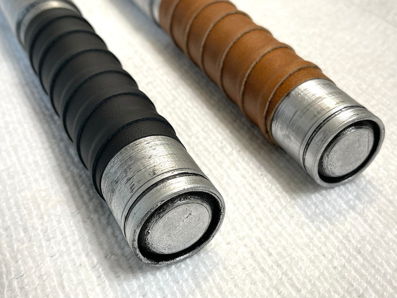 K&ouml;nnte beinhalten: Zwei Lichtschwerter mit Griffen in verschiedenen Farben. Ein Griff hat einen schwarzen Ledergriff und der andere einen braunen Ledergriff. Beide Griffe haben eine silberne Metalloberfl&auml;che.