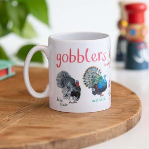 Puede incluir: Taza de cerámica blanca con la palabra "gobblers" en escritura rosa. La taza presenta ilustraciones de dos pavos, etiquetados como "blue slate" y "occellated". La taza tiene un asa blanca y está sobre una superficie de madera.