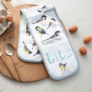 Op de afbeelding: Een paar blauwe en witte ovenwantjes met een speels ontwerp met verschillende soorten vogels, waaronder een blauwe mees, een koolmees, een wilgmees en een staartmees. De wantjes zijn bedrukt met het woord "tits" in een groot, speels lettertype.