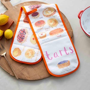 May include: A pair of white oven mitts with orange trim featuring illustrations of different types of tarts with names like "Jam", "Caramel", "Bakewell", and "Tatin". The mitts have the word "tarts" printed in pink letters.