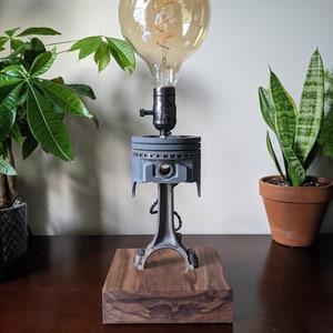 May include: A unique desk lamp featuring an Edison-style light bulb. The lamp's base is a rectangular block of dark wood, supporting a repurposed engine piston and connecting rod. The bulb is large and round, with a visible filament.