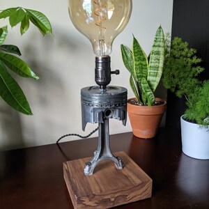 May include: A table lamp made from a repurposed engine piston and a wooden base. The lamp has a large, round, clear glass bulb with a coiled filament.