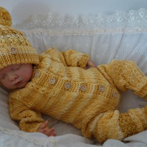 Peut inclure: Ensemble de bébé tricoté jaune, comprenant un bonnet à pompon, une barboteuse boutonnée et des chaussons assortis. L'ensemble est en fil doux et est conçu pour un nouveau-né. Le bébé est dans un berceau blanc bordé de dentelle.